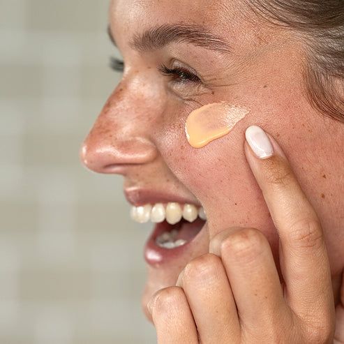 Person trägt Creme auf die Wange auf. Creme ist gelblich. Person lächelt.