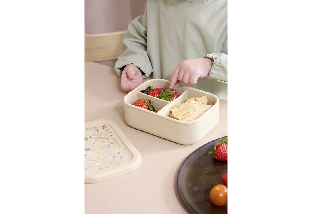 Kind greift in beige Brotdose mit Erdbeeren und Cracker. Deckel daneben. Aufteilung in drei Fächer. Auf Tisch.