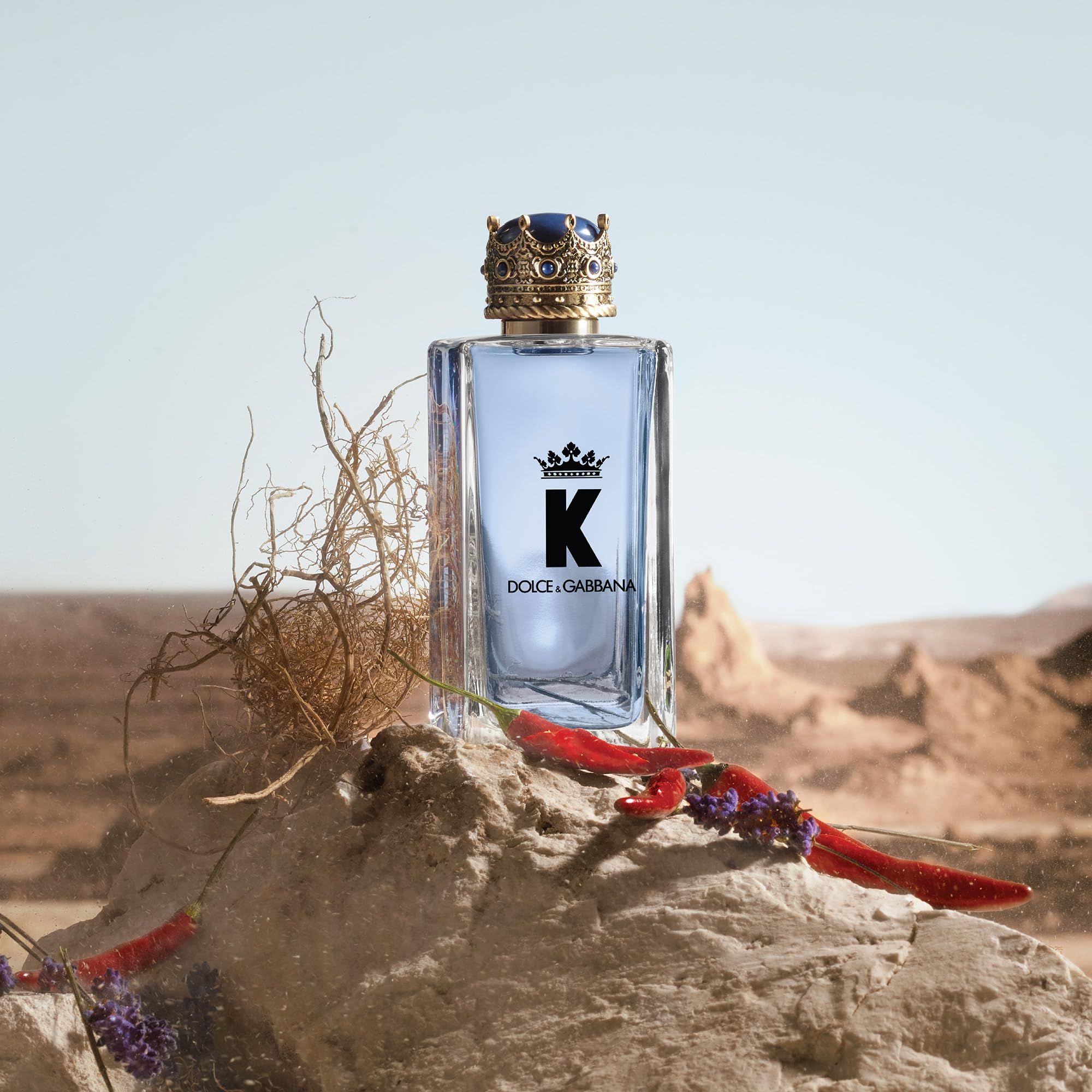 K by Dolce & Gabbana Flakon auf einem Felsen. Flakon mit Kronenkappe. Rote Paprika und trockene Pflanzen umgeben den Flakon.