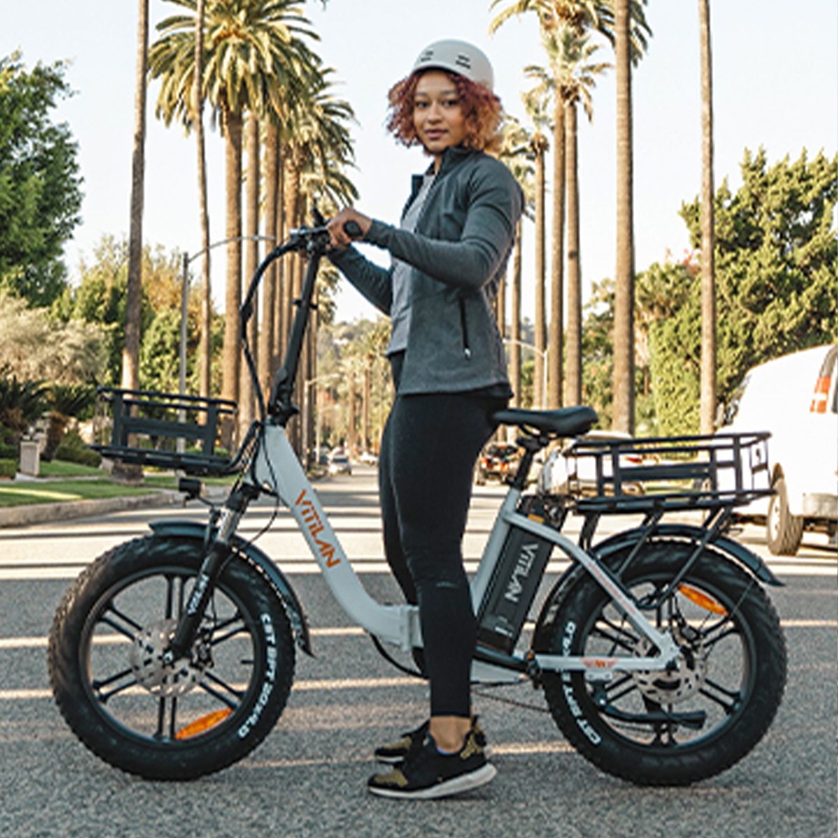 Person steht neben weißem Vitilan Elektrofahrrad. Sie trägt Helm und Sportkleidung. Im Hintergrund Palmen.
