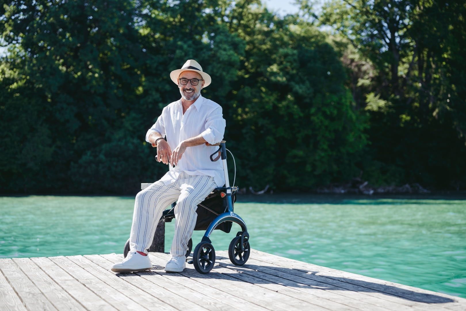 Älterer Mann sitzt auf einem blauen Rollator auf einem Steg. Er trägt Hut, Hemd und Hose. Hintergrund: See und Bäume.