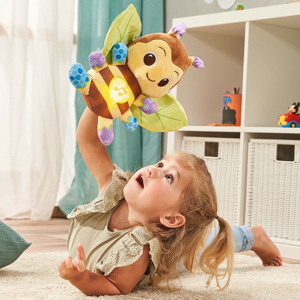 Kind hält Vtech Musik-Kuschelbiene hoch. Die Biene ist braun, gelb und grün. Kind liegt auf dem Boden.