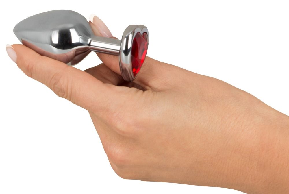 Hand hält Analplug mit herzförmigem, rotem Schmuckstein. Silberfarbenes Metall, herzförmiger Sockel.