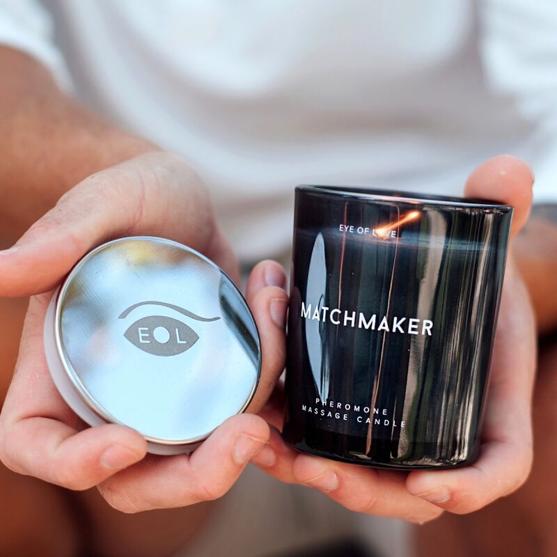Person hält eine schwarze Kerze und einen silbernen Deckel. Aufschrift: EYE OF LOVE, MATCHMAKER, PHEROMONE MASSAGE CANDLE.