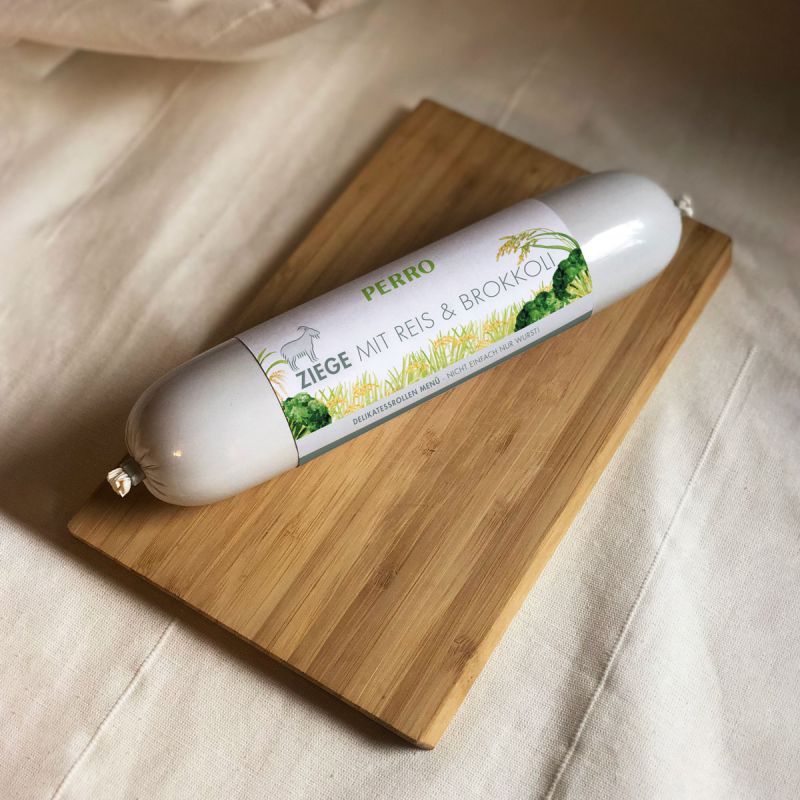 Wurstrolle auf Holzbrett. Etikett: PERRO, ZIEGE MIT REIS & BROKKOLI. Produkt für Hunde.