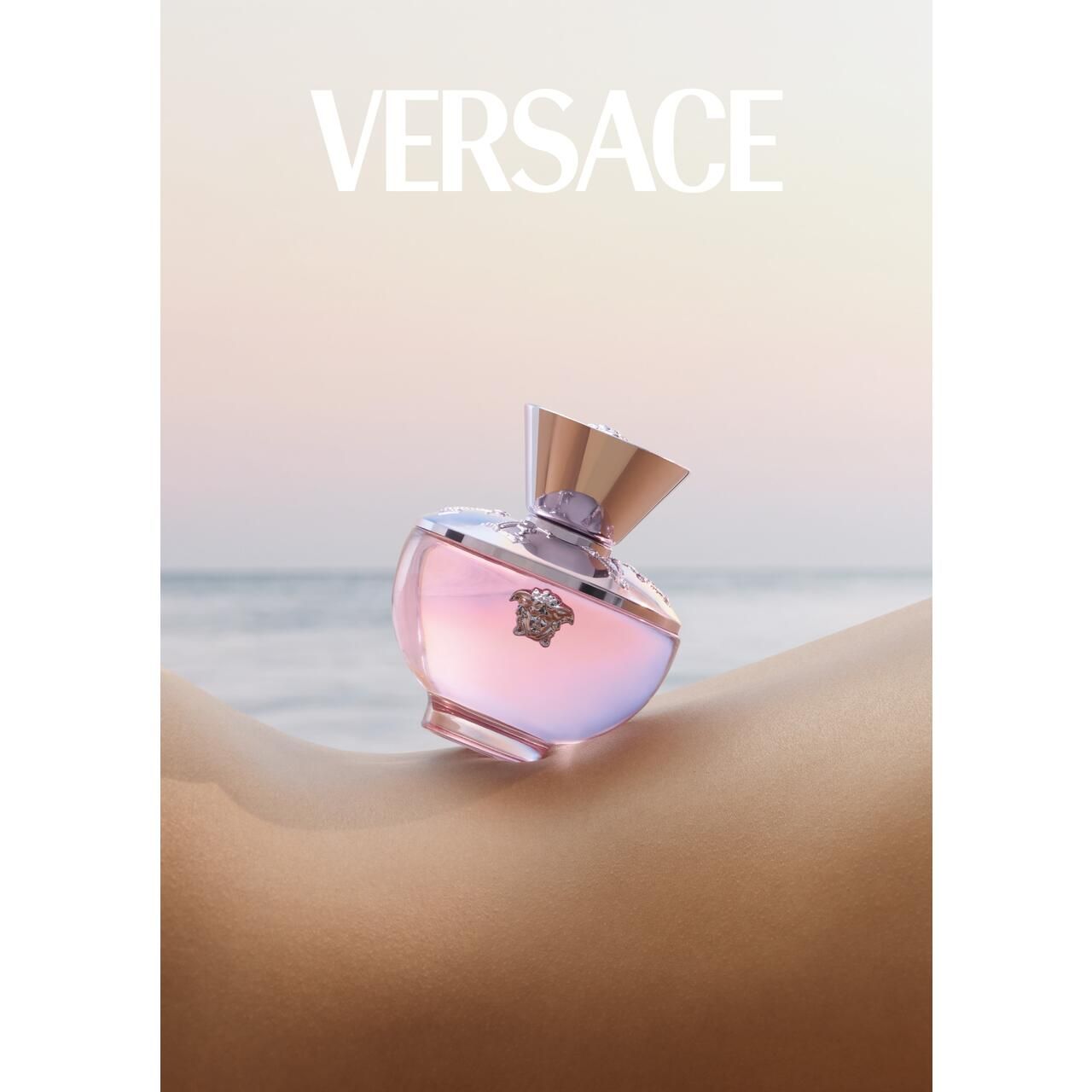 Versace Dylan Blush Pink Eau de Parfum Flakon auf Haut. Flakon mit rosa Glas, silbernem Deckel und Medusa-Logo. Hintergrund: Haut und Meer.