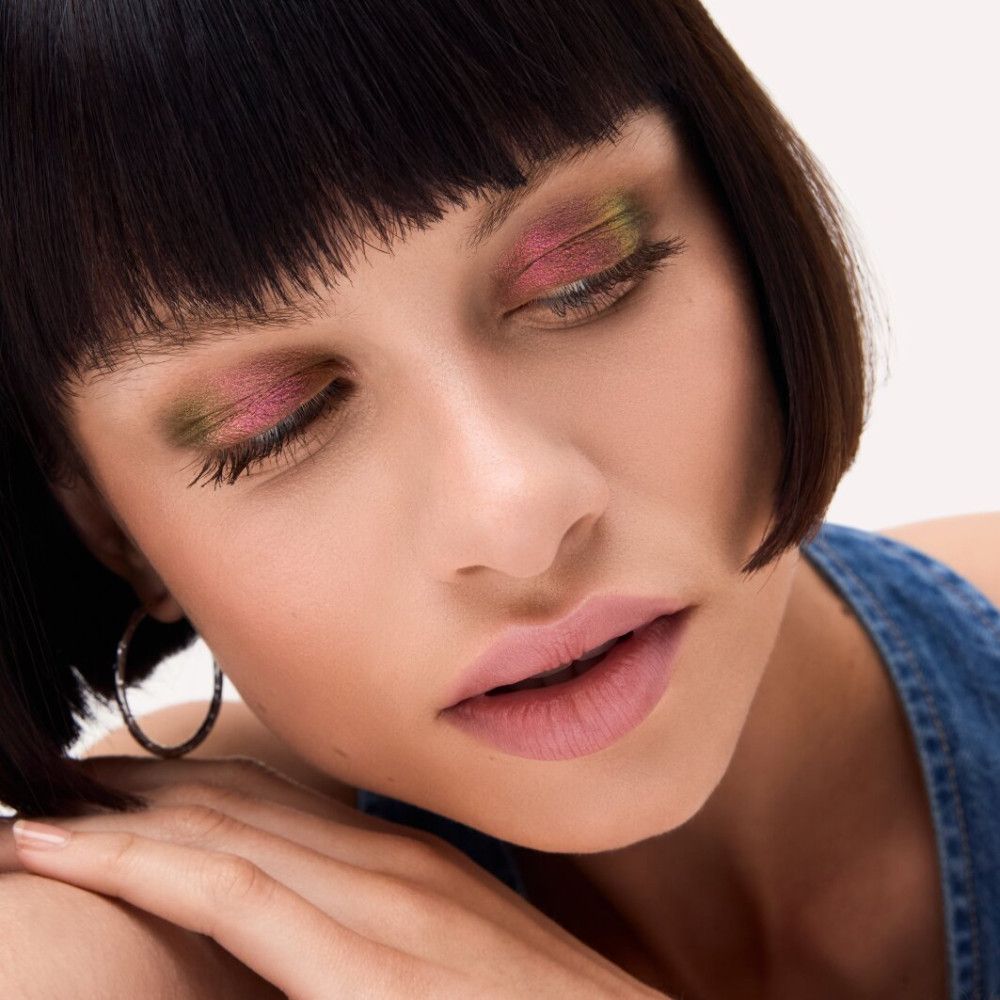 Nahaufnahme eines Gesichts mit geschminkten Augen. Rosa und grüne Lidschatten. Die Frau hat kurze, dunkle Haare und trägt einen blauen Jeans-Overall.