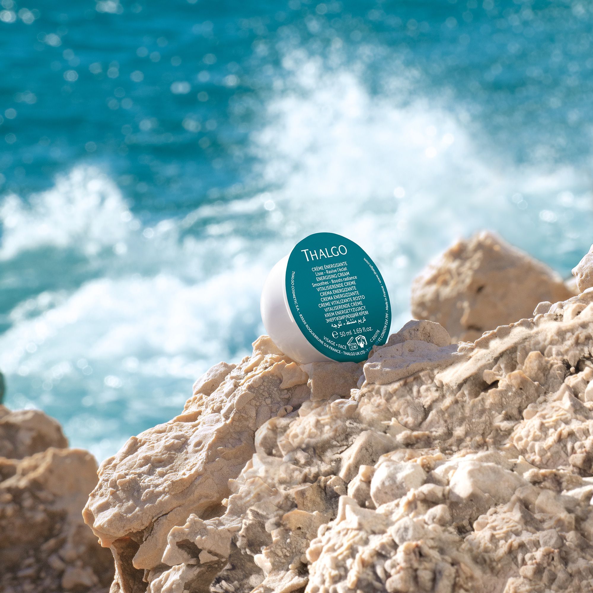 Weiße Creme-Verpackung mit blauem Deckel auf Felsen am Meer. Aufschrift: THALGO, Creme Energisante.
