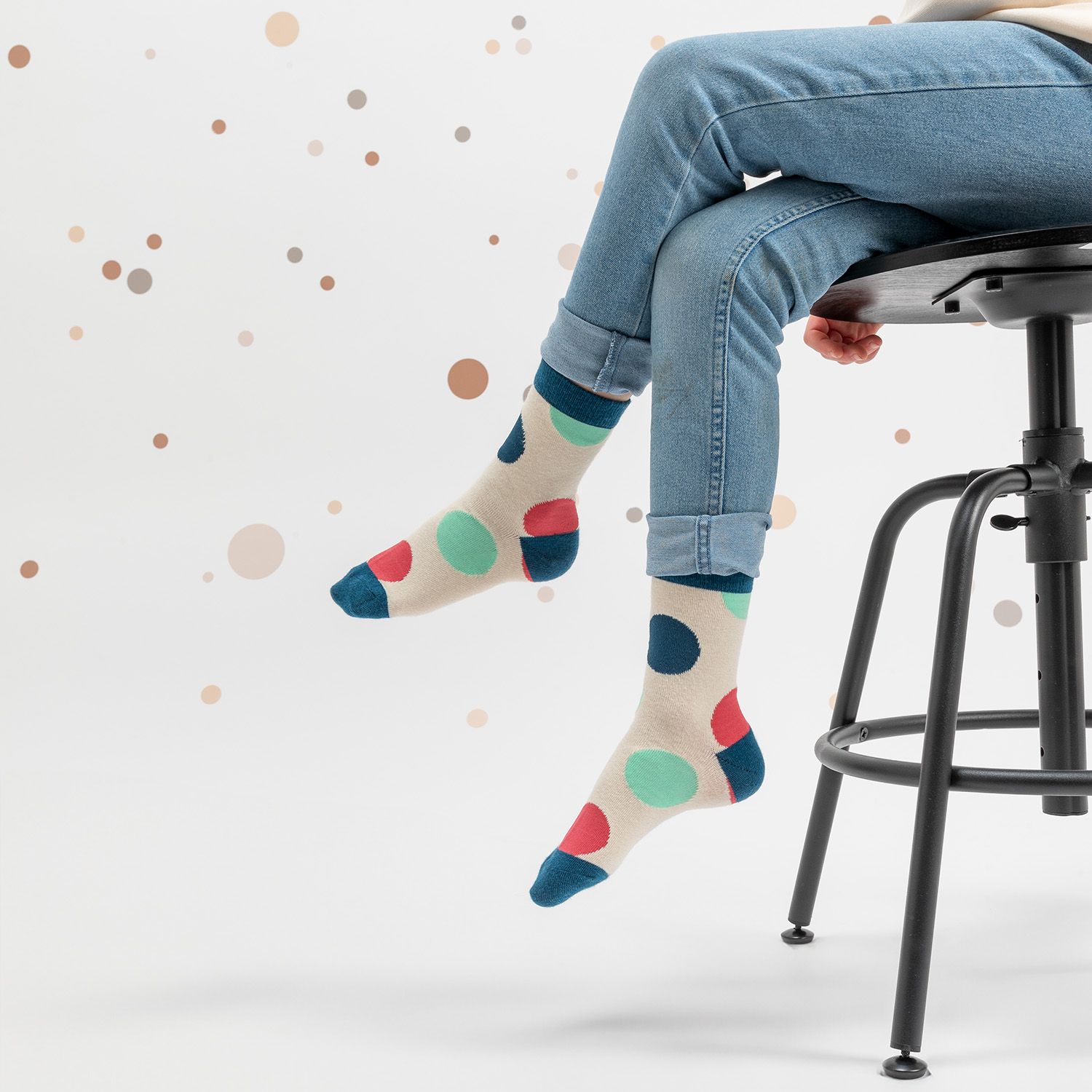 Beige Socken mit bunten Punkten. Kind sitzt auf einem Hocker. Hintergrund: weiß mit Punkten.