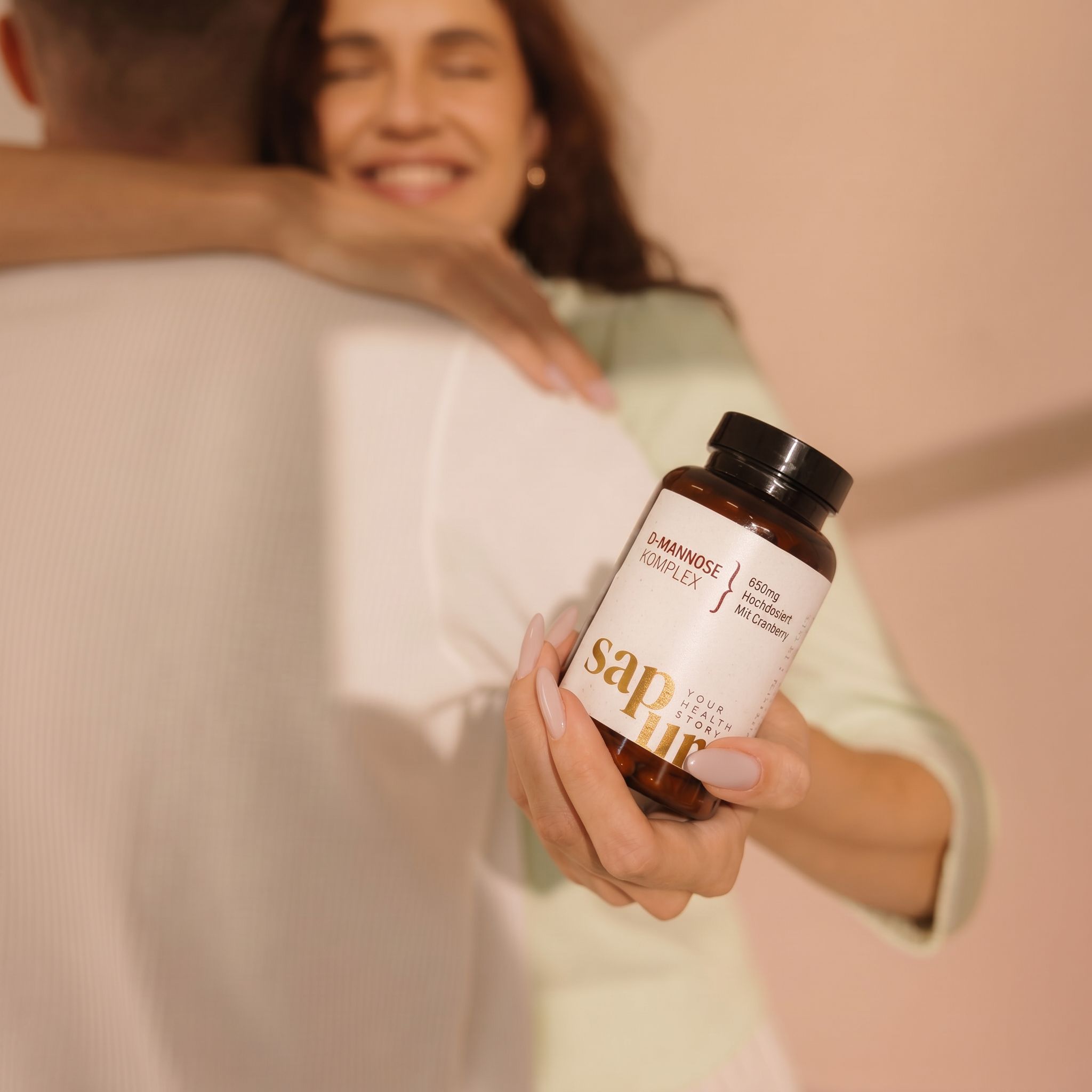 Person hält eine braune Flasche mit Etikett. Aufschrift: D-Mannose Komplex+, 650mg, Mit Cranberry, sapura health.
