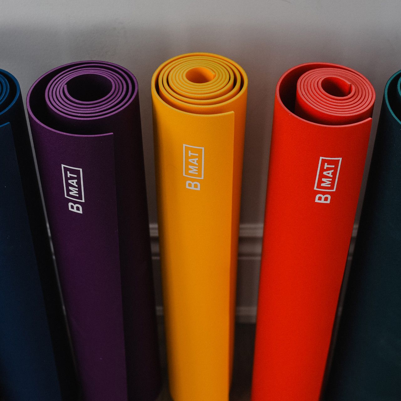 Fünf gerollte Yogamatten in verschiedenen Farben. Logo "B MAT" sichtbar.