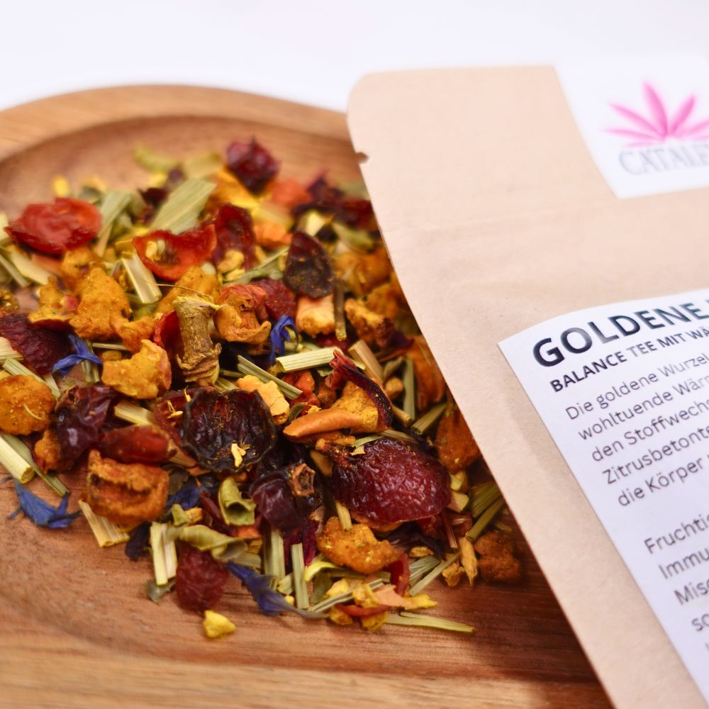 Tee-Mischung auf Holzschale neben offener Verpackung. Aufdruck 'GOLDENE HARMONIE' und Logo. Kräuter und Früchte sichtbar.