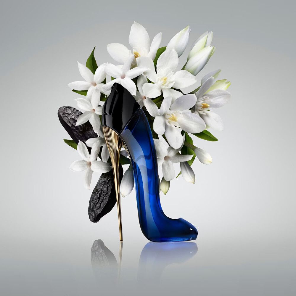 Blauer High Heel-Flakon mit goldenem Absatz, umgeben von weißen Blüten und dunklen Bohnen. Auf hellem Hintergrund.
