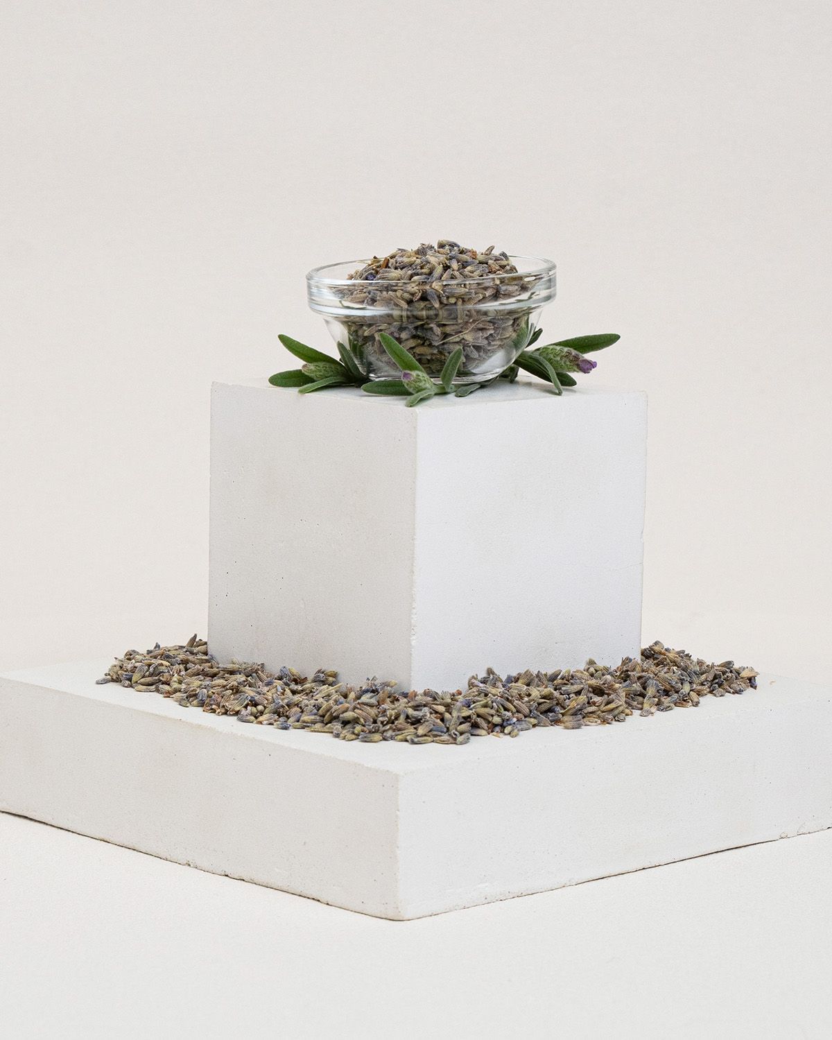 Kleine Glasschale mit Lavendelblüten auf einem Podest. Umgeben von Blüten und Zweigen.