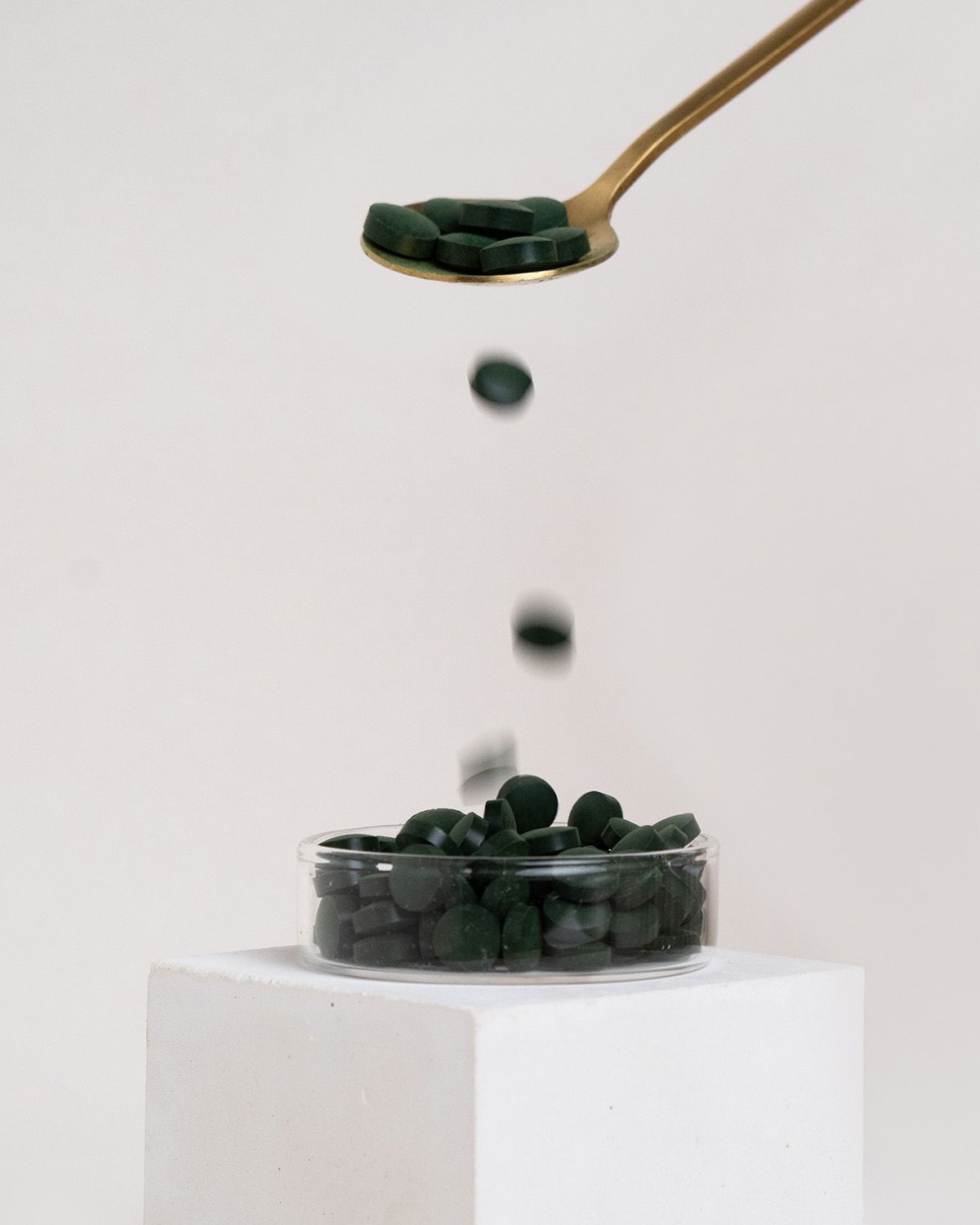 Grüne Tabletten fallen von einem goldenen Löffel in eine Glasschale. Die Tabletten sind rund und flach.