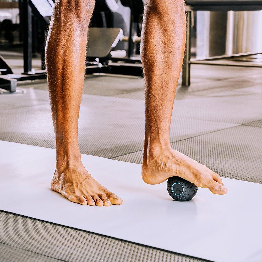 Füße auf einer Matte, ein schwarzer Massageball unter dem Fuß. Fitnessstudio-Umgebung.