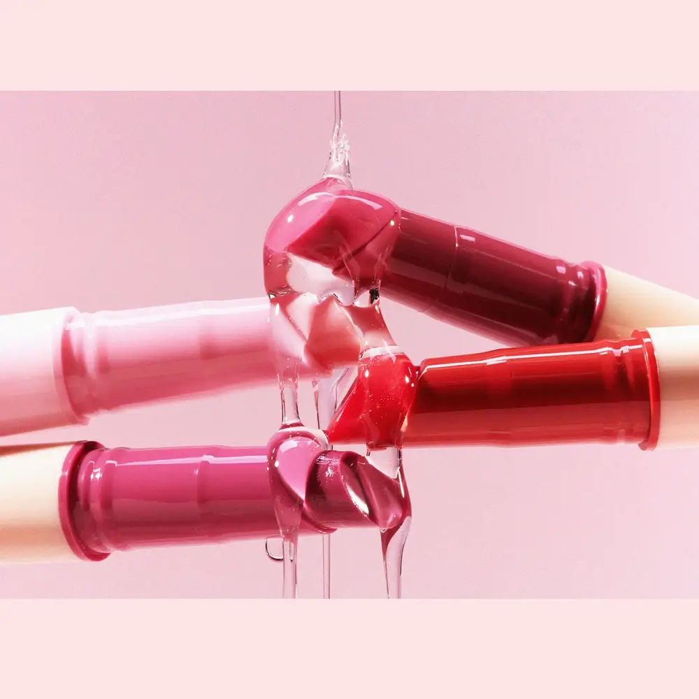 Drei Lipgloss-Stifte, aus denen Lipgloss tropft. Farben: Rosa, Rot, Pflaume. Hintergrund: rosa.