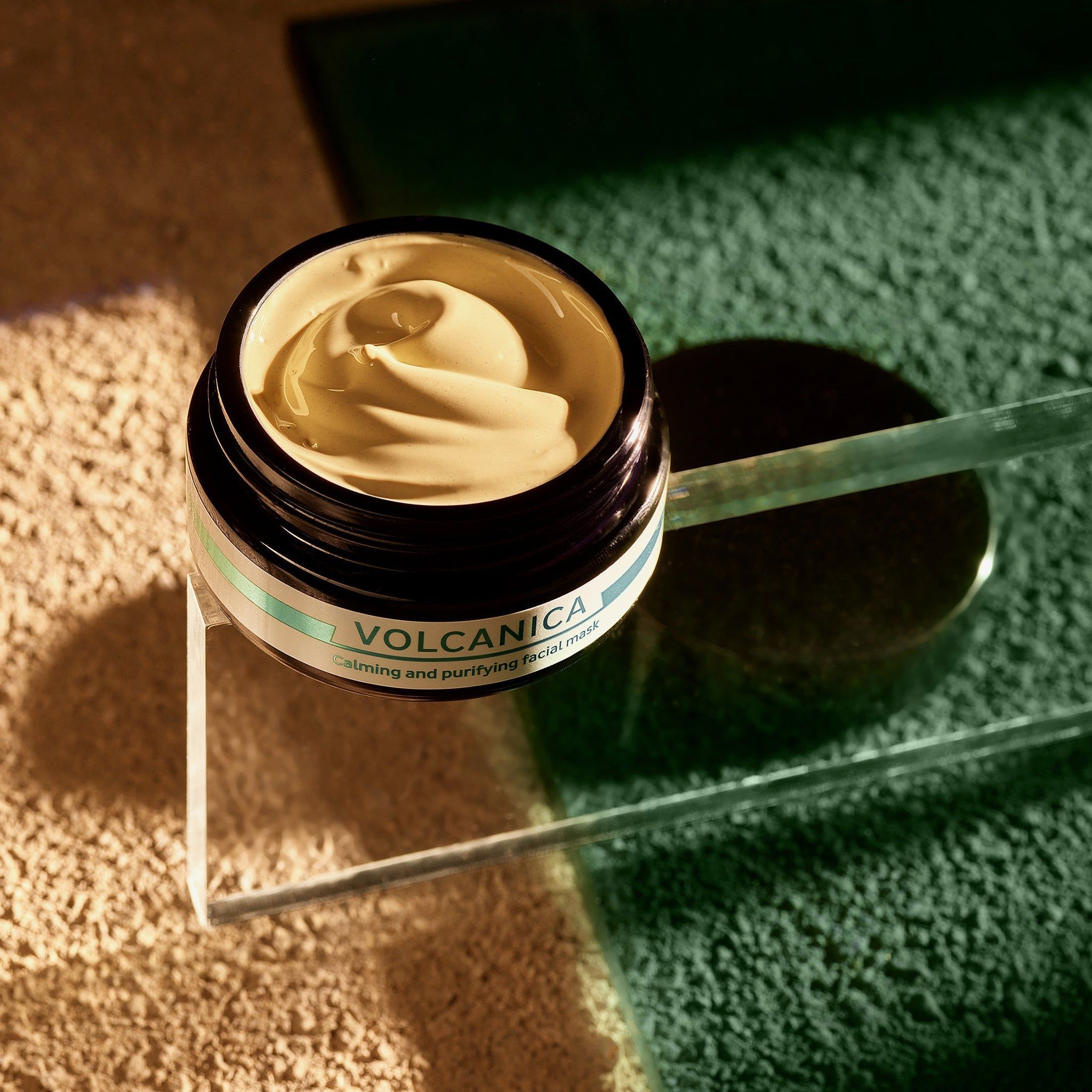 Geöffnetes Tiegel mit cremiger Maske. Aufschrift: VOLCANICA, Calming and purifying facial mask. Auf Glasplatte.