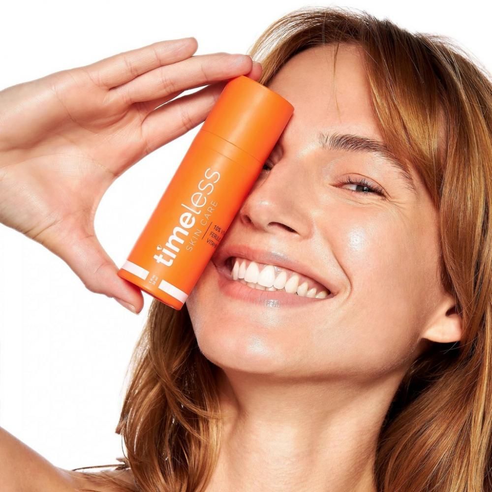 Frau hält eine orangefarbene Flasche. Aufschrift: Timeless Skin Care. Produkt: 10% Vitamin C, Ferulasäure, Vitamin E.