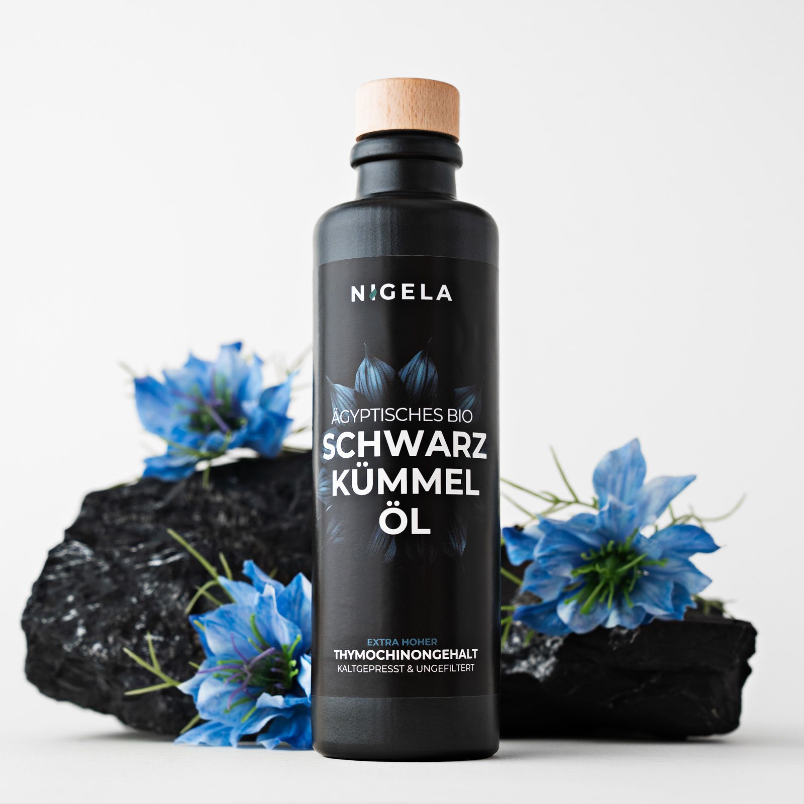 Schwarze Flasche mit Holzverschluss. Aufschrift: Nigela, Ägyptisches Bio Schwarzkümmel Öl. Dekoration: blaue Blüten.