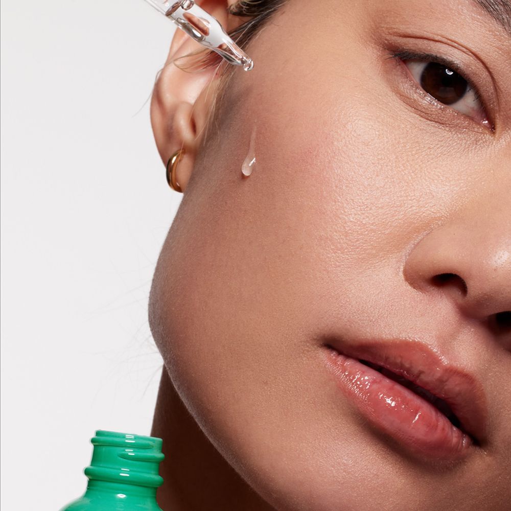 Nahaufnahme einer Frau. Ein Tropfen Serum fällt auf die Haut. Grüne Flasche im Hintergrund.