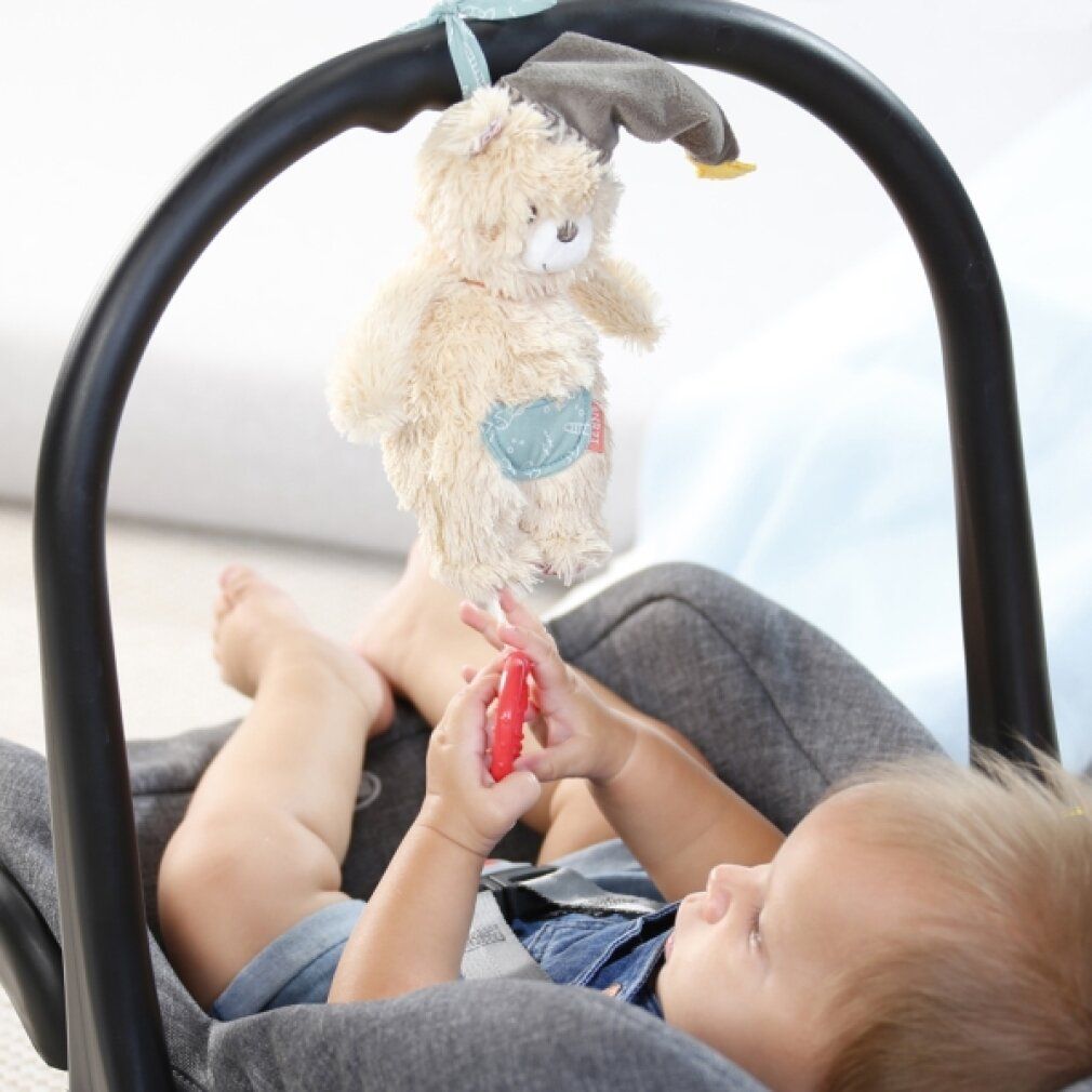 Beige Bär-Spieluhr an einem schwarzen Bogen über einem Baby. Das Baby liegt in einem Autositz und hält ein rotes Spielzeug.