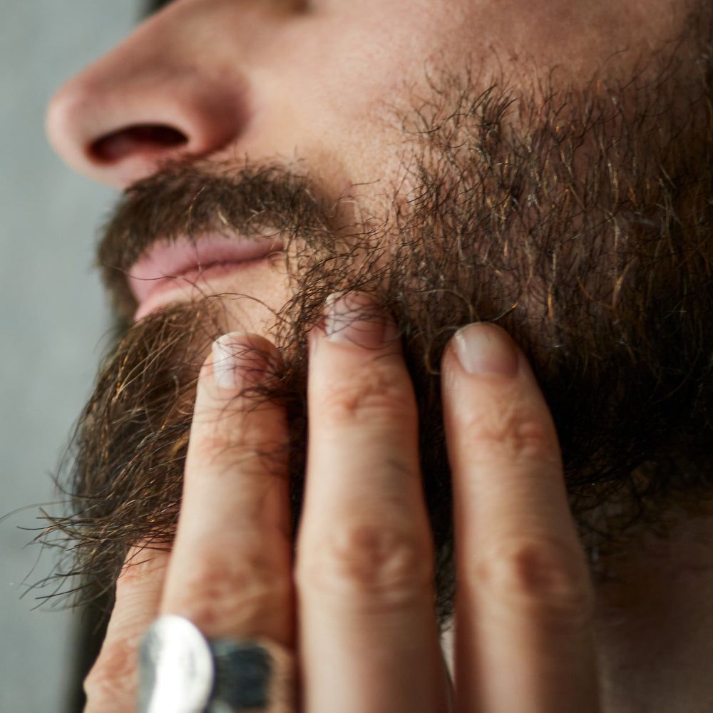 Nahaufnahme eines Mannes, der seinen Bart berührt. Der Bart ist braun und buschig. Ein silberner Ring ist am Finger zu sehen.