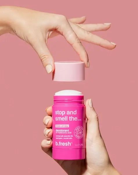 Rosa Deodorant-Stick, geöffnet. Hände halten den Stick und den Deckel. Rosa Verpackung, weißer Deodorant-Kopf. Text: B.fresh, Stop and smell the...
