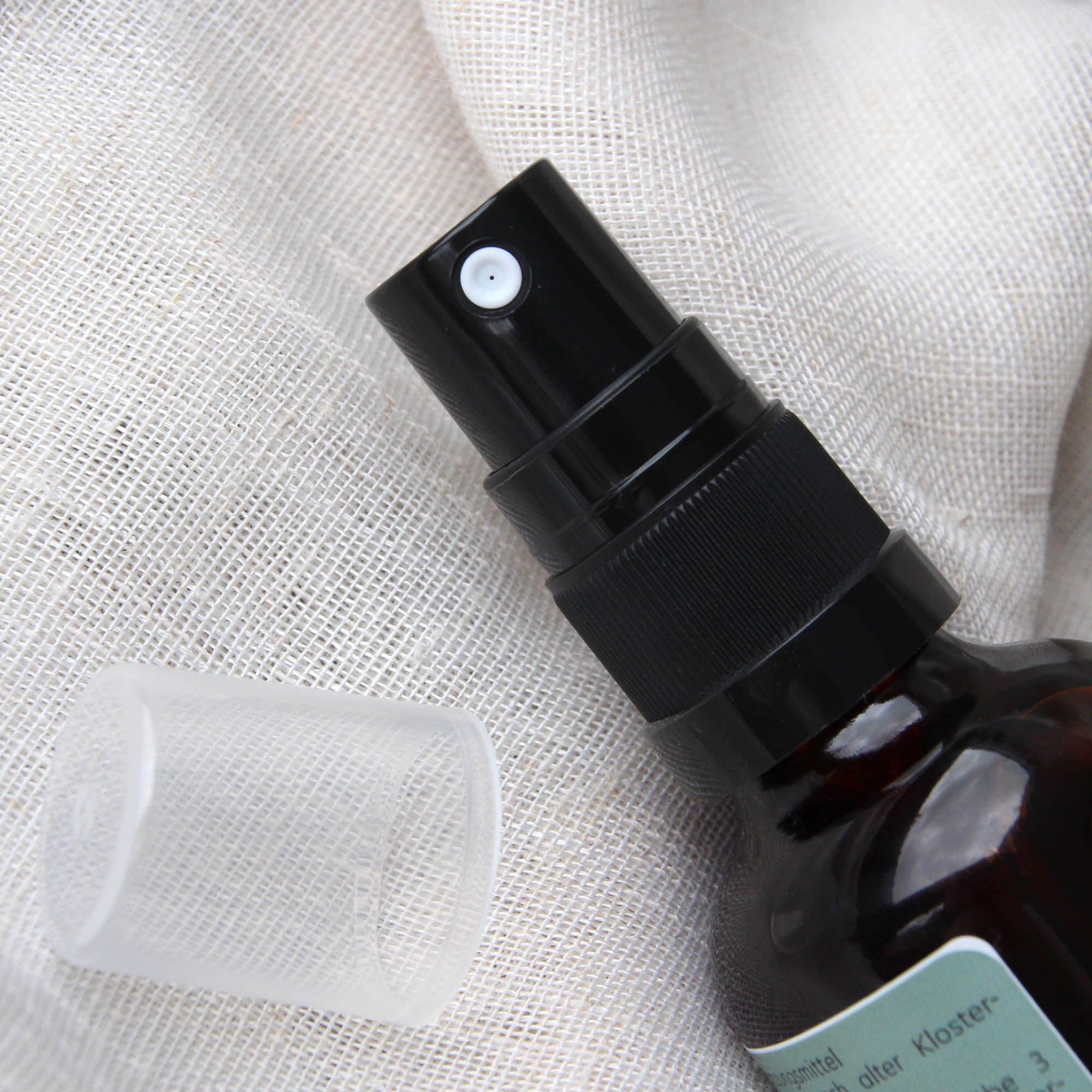 Braune Glasflasche mit schwarzem Sprühkopf und transparenter Kappe. Auf hellem Stoff platziert.
