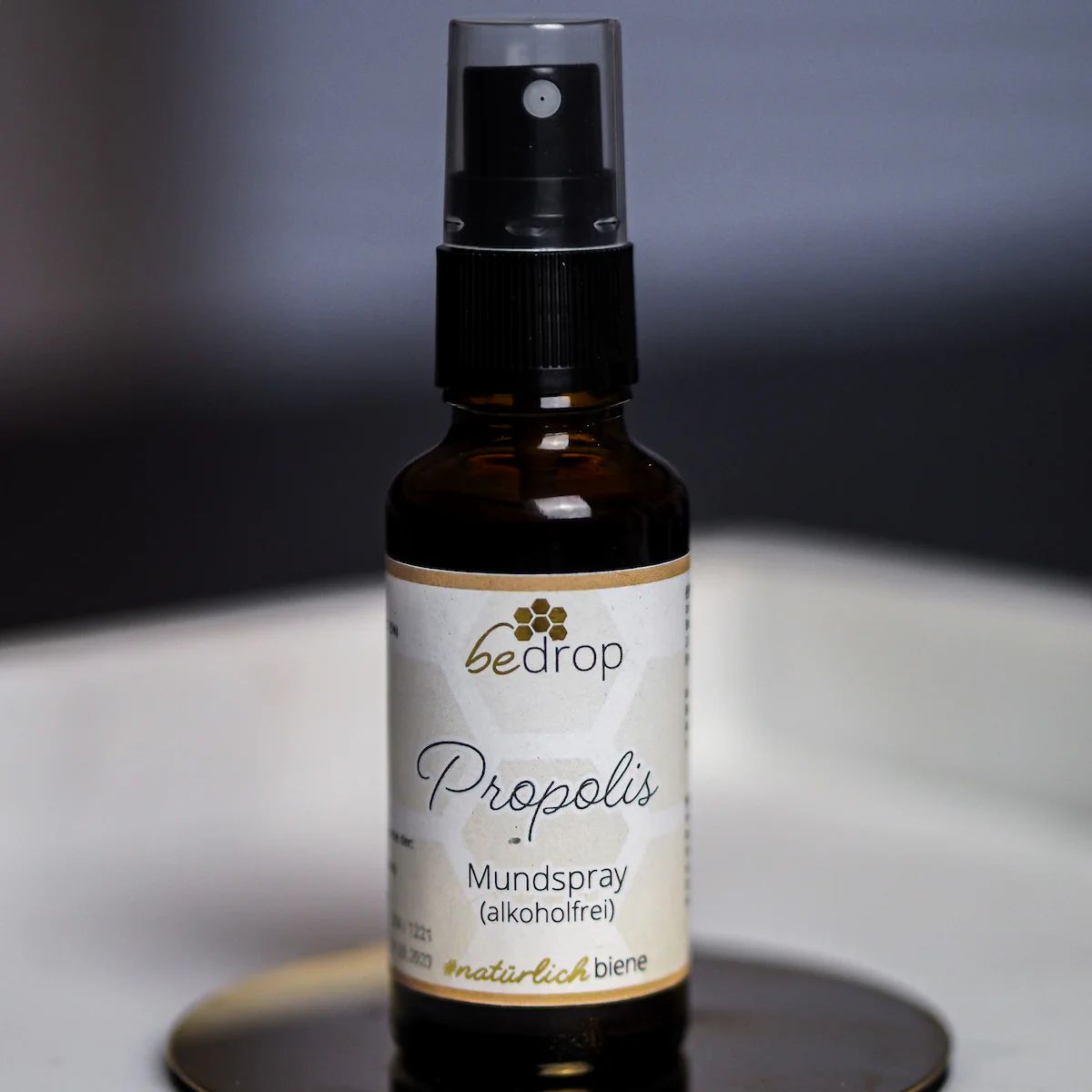 Braune Glasflasche mit schwarzem Sprühkopf. Aufschrift: bedrop Propolis Mundspray. Steht auf einer runden Unterlage.