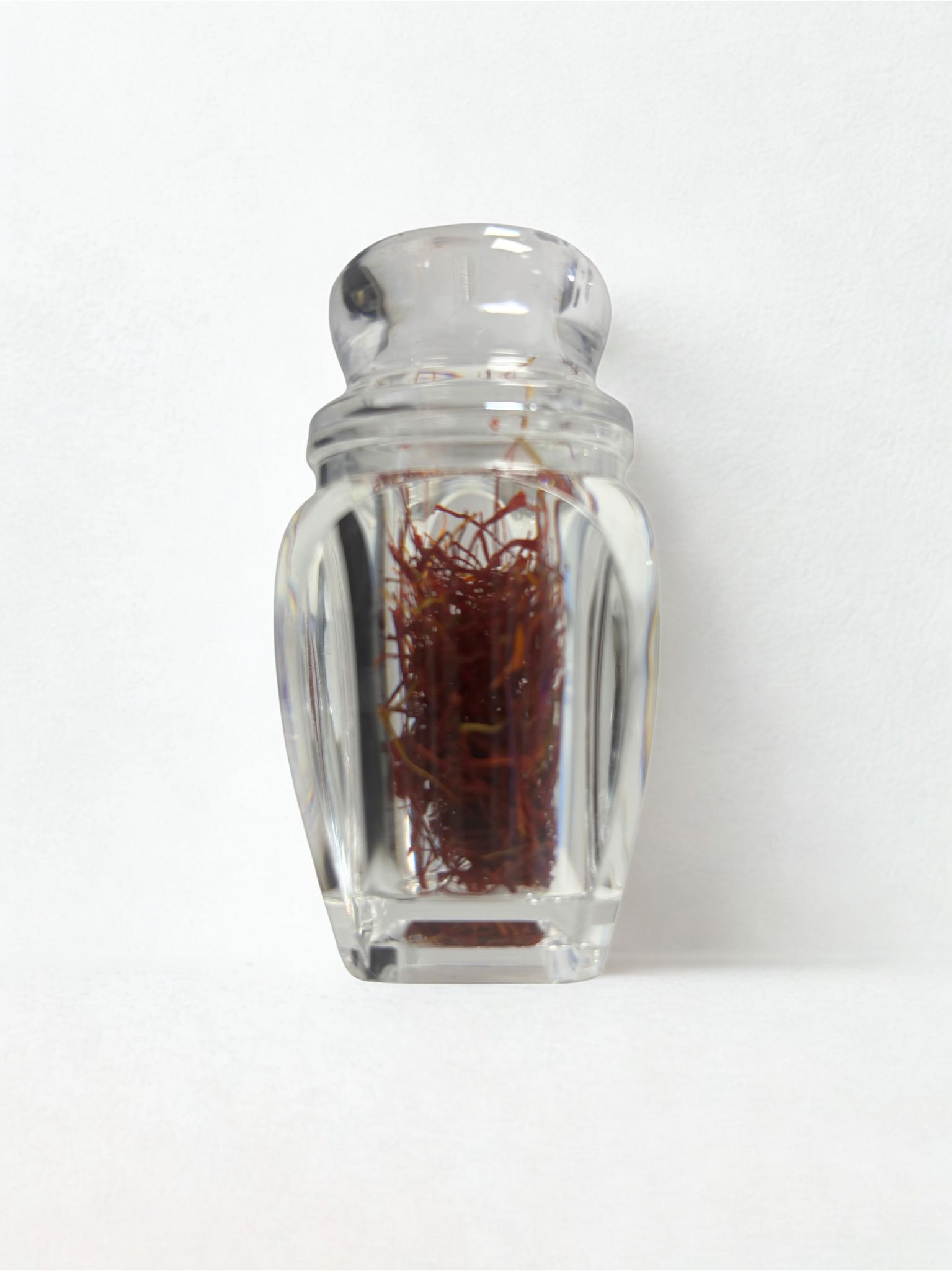 Glasbehälter mit Safranfäden. Klarer, quadratischer Behälter mit Deckel. Rote Safranfäden sichtbar.