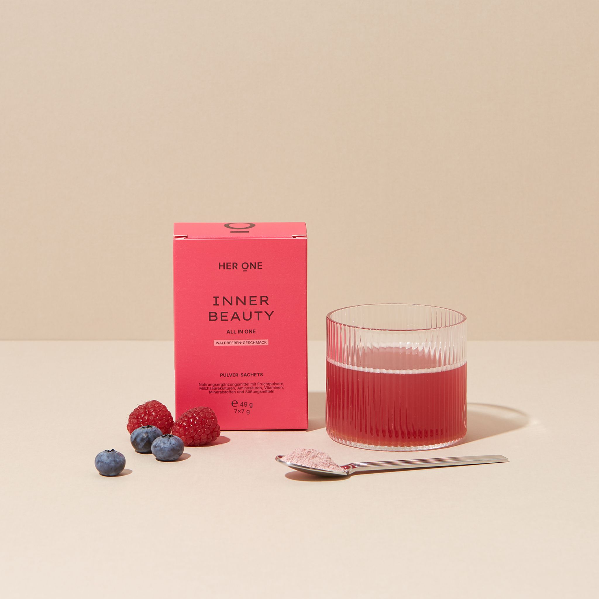 Produktverpackung neben Glas mit Getränk, Löffel mit Pulver, Blaubeeren und Himbeeren. Rosa Verpackung mit Text.