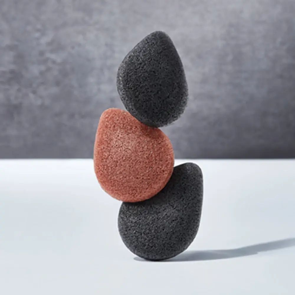 Drei Konjak-Schwämme, zwei grau, einer rotbraun, in Balance gestapelt. Hintergrund: weiß und grau.
