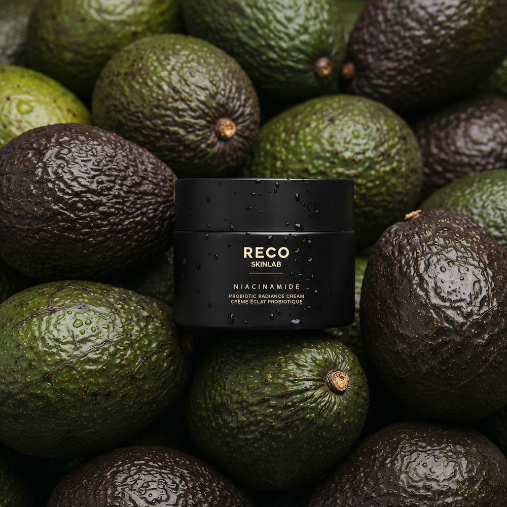 Schwarzer Tiegel auf Avocados. Aufschrift: RECO SKINLAB, Niacinamide Probiotic Radiance Cream. Grüner Hintergrund.