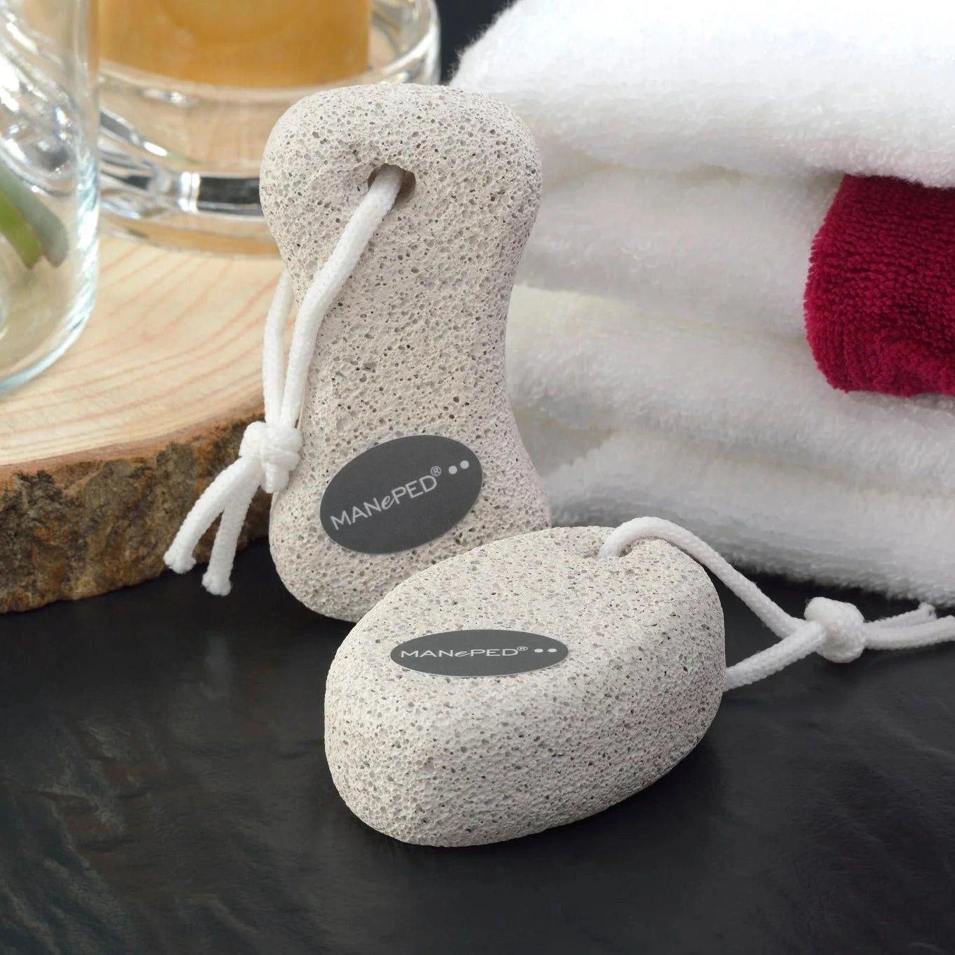Zwei Bimssteine mit weißer Schnur. MANePED-Logo. Auf schwarzem Untergrund, Handtücher im Hintergrund.