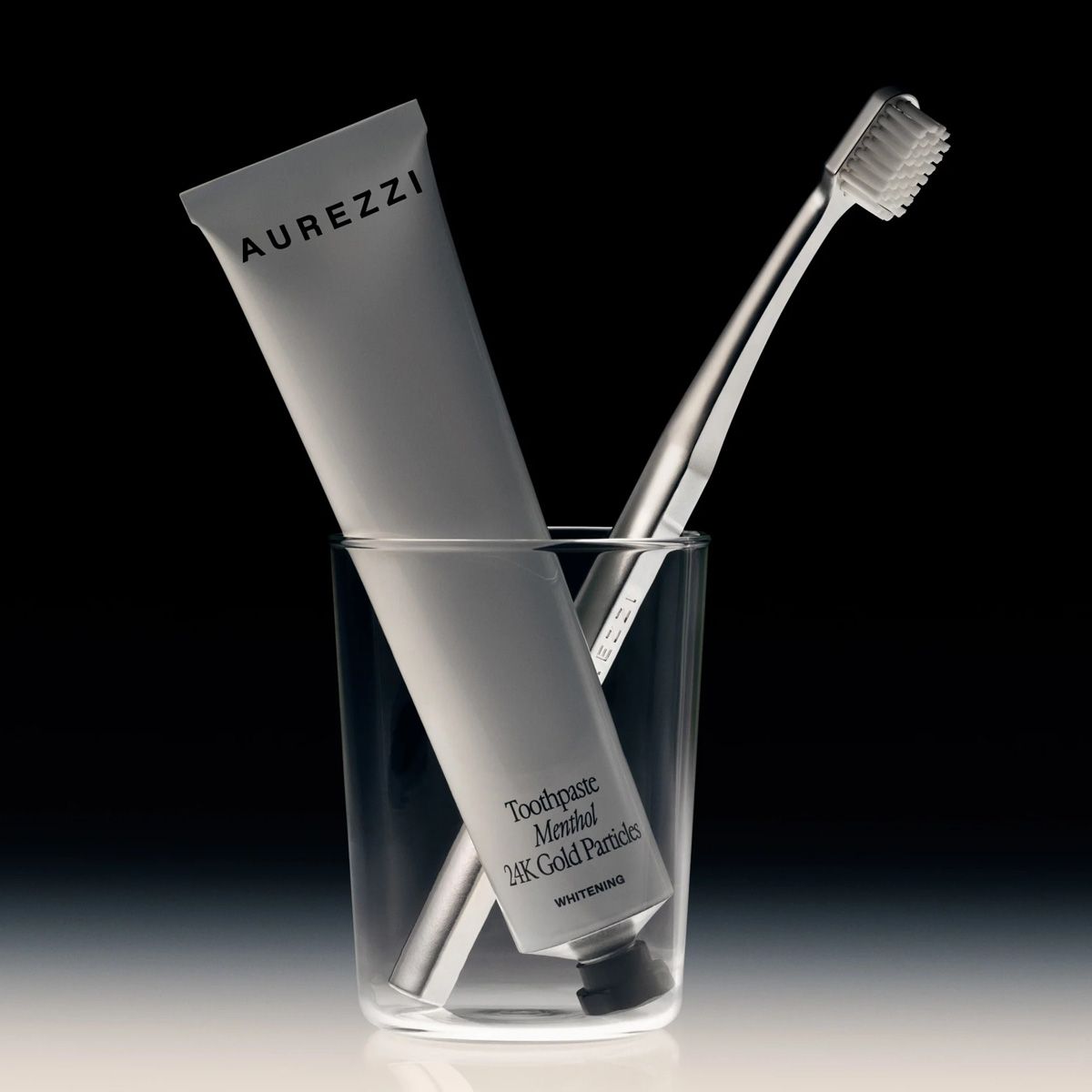Zahnpasta und Zahnbürste in Glas. Zahnpasta-Tube mit Aufschrift AUREZZI. Zahnbürste mit silberfarbenem Griff.