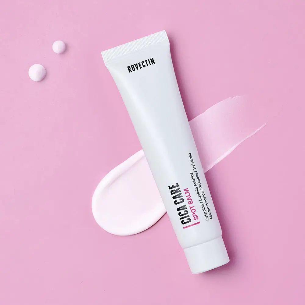 Weiße Tube mit Produkt. Aufschrift: Rovectin, Cica Care Spot Balm. Rosa Hintergrund mit Produkt-Ausstreichungen.