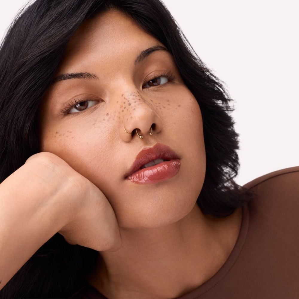 Porträt einer Frau mit Lipgloss. Farbe: orange-braun. Sie stützt ihr Kinn auf ihre Hand.