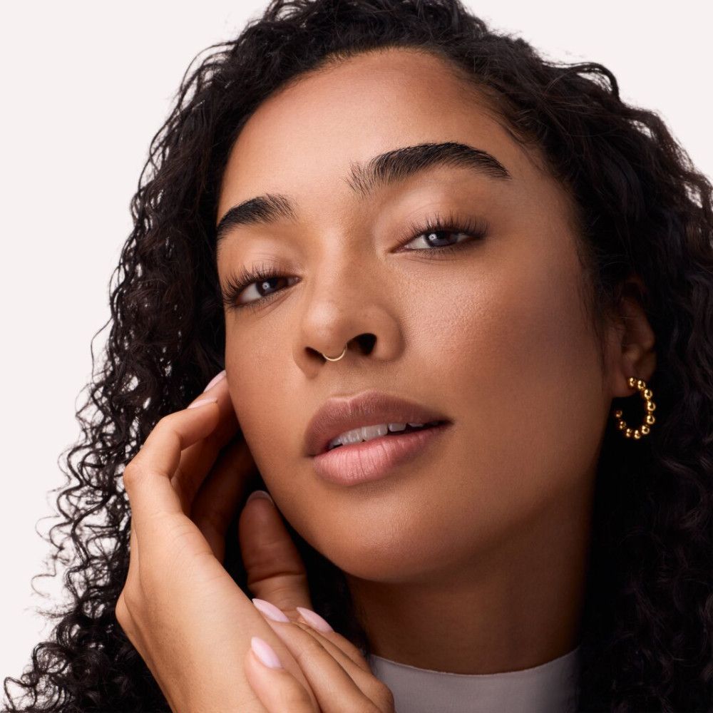 Nahaufnahme eines Gesichts. Gebräunte Haut, dunkle Locken, Nasenring, goldener Ohrring. Hand berührt Wange. Natürliches Licht.