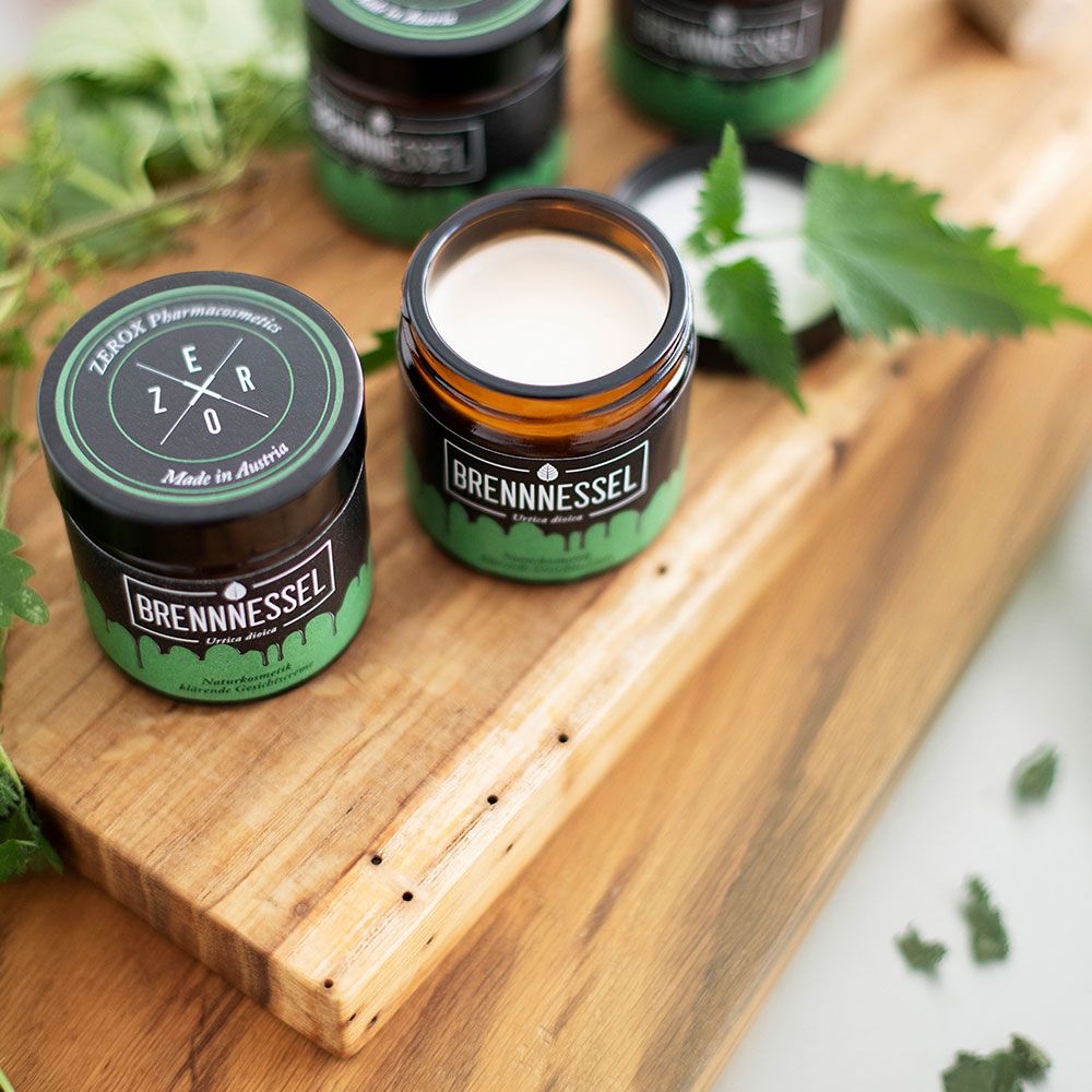 Mehrere Gläser mit schwarzem Deckel und grünen Etiketten. Ein Glas geöffnet, Creme sichtbar. Auf Holzbrett mit Brennnesselblättern.