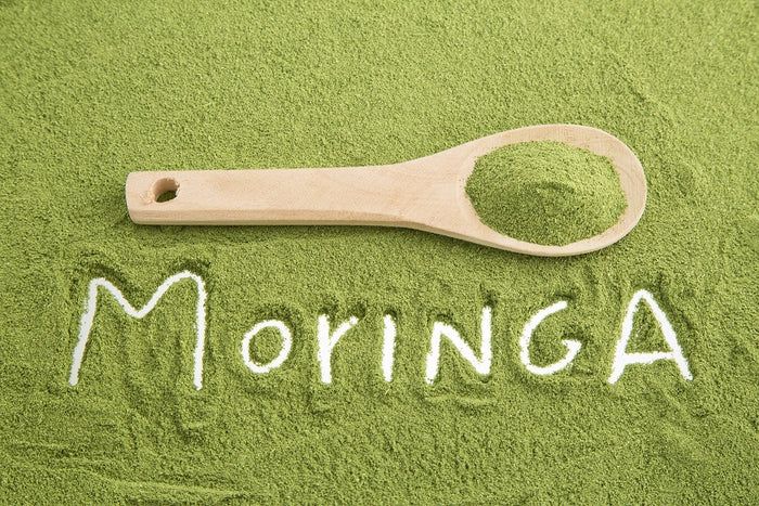 Grünes Pulver mit dem Wort "MORINGA" und einem Holzlöffel.