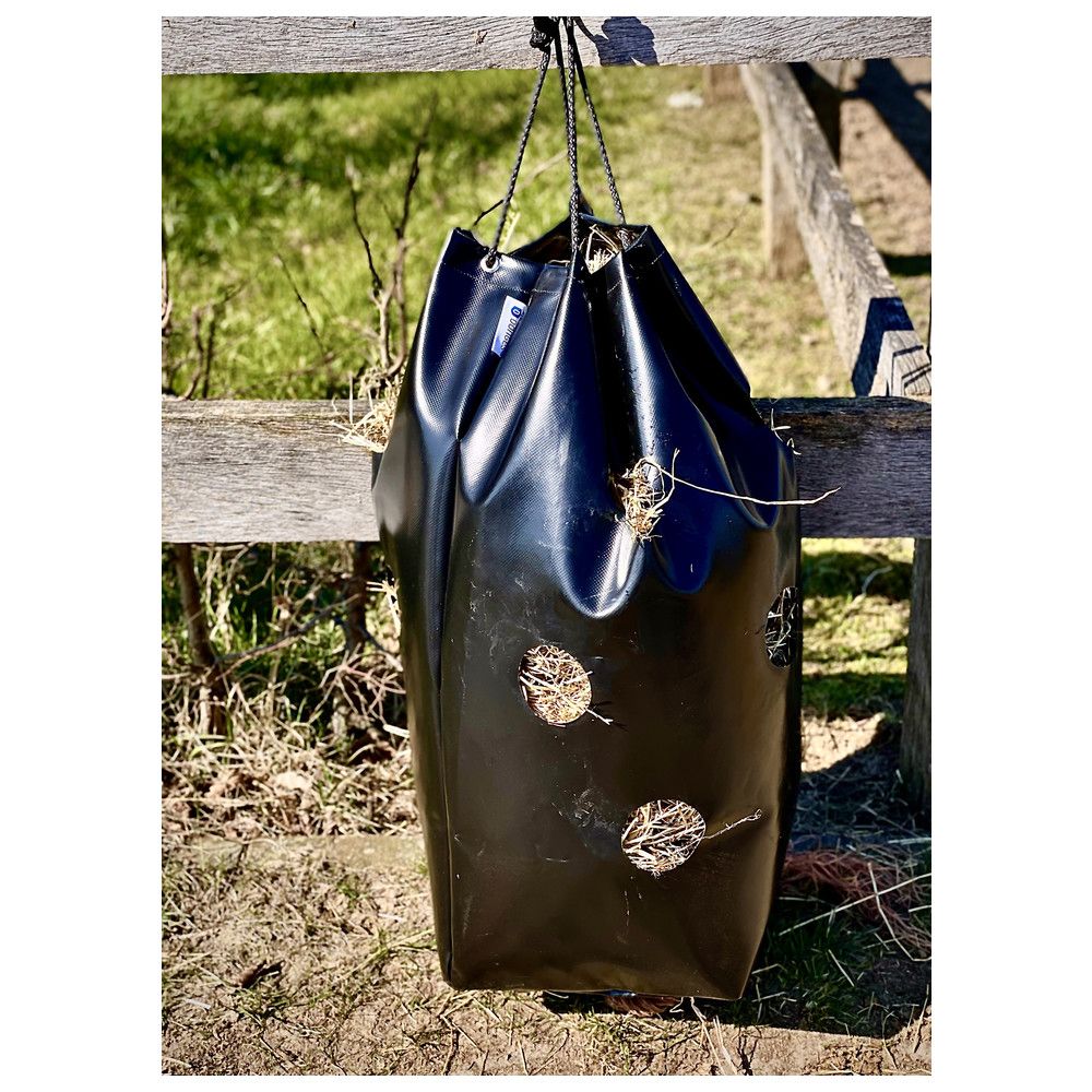 Schwarzer Heusack hängt an einem Zaun. Lochöffnungen für das Heu.