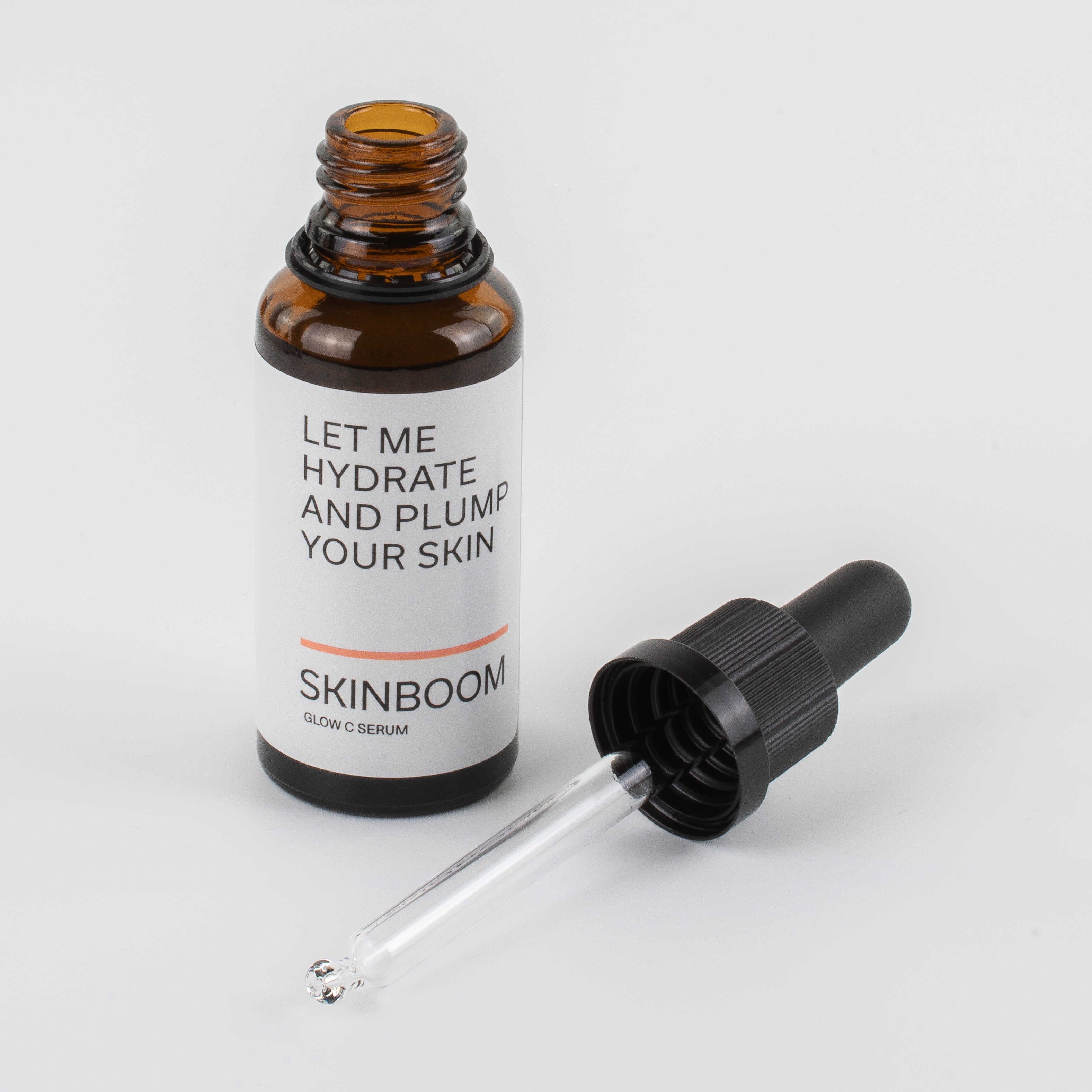 Geöffnete braune Glasflasche mit Pipette daneben. Etikett mit Text: LET ME HYDRATE AND PLUMP YOUR SKIN. SKINBOOM Glow C Serum.
