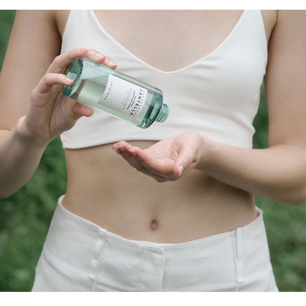 Person gießt grüne Flüssigkeit aus einer Flasche in die Handfläche. Trägt weißes Top und Hose. Hintergrund: Natur.