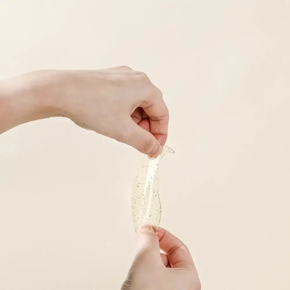 Hände halten eine transparente Augen-Hydrogel-Maske. Die Maske hat eine geschwungene Form und schwarze Punkte.