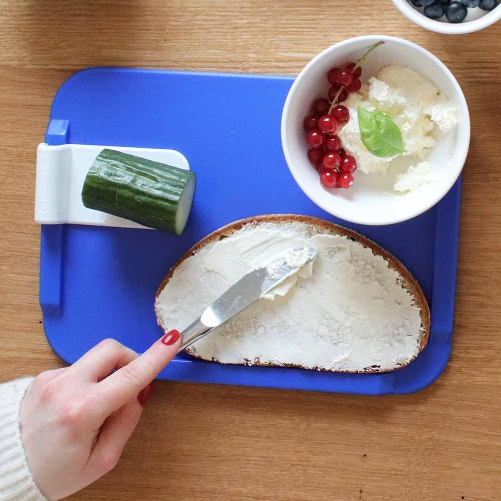 Blaues Schneidebrett mit Gurkenscheibe und Schale mit Beeren. Hand bestreicht Brot mit Frischkäse.