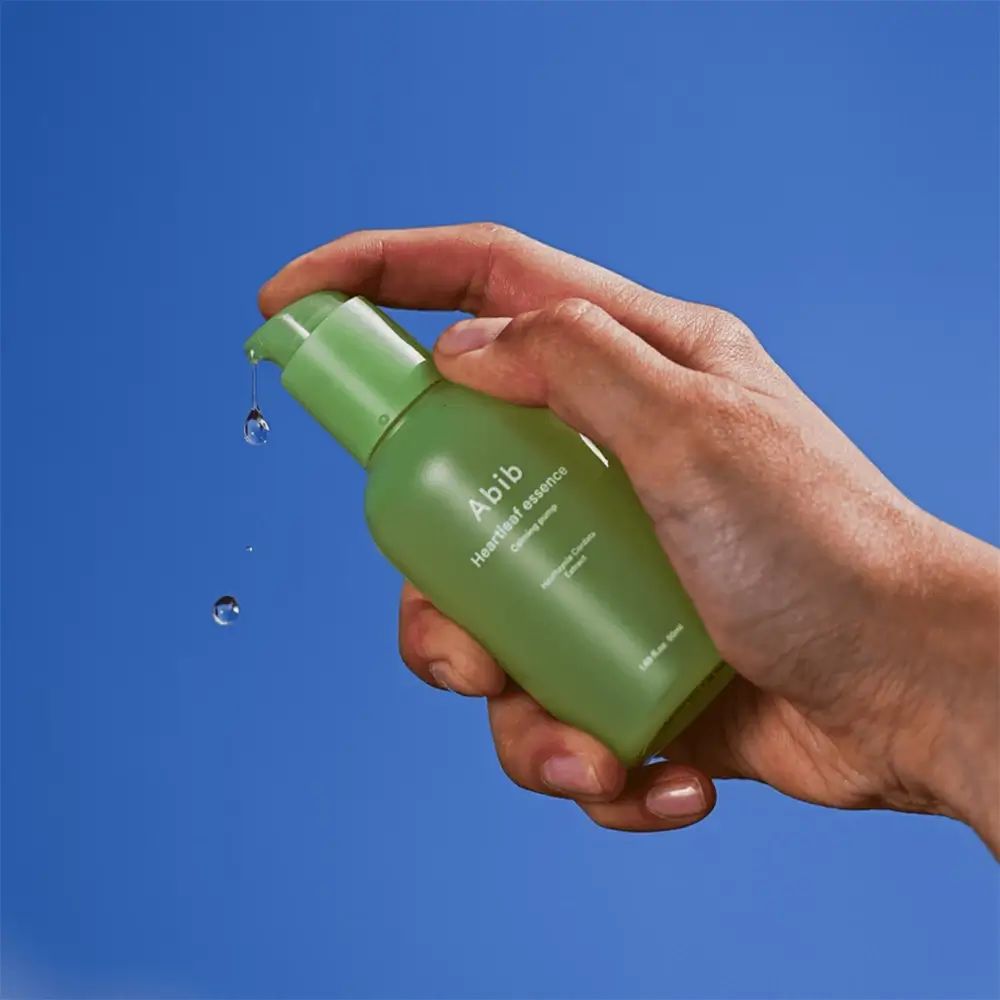 Grüne Flasche mit Pumpe, gehalten von einer Hand. Tropfen fallen. Aufschrift: Abib Heartleaf essence Calming pump. Blauer Hintergrund.