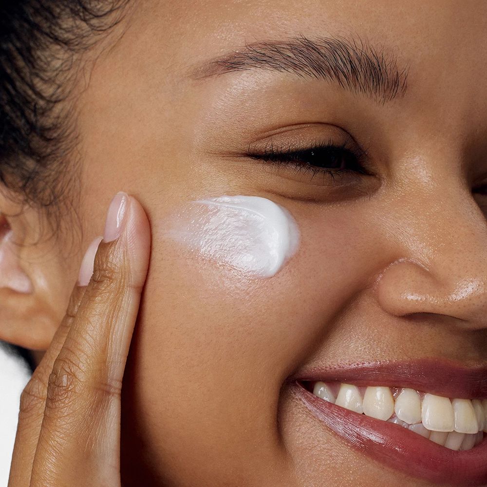Person trägt Creme auf die Wange auf. Weiße Creme auf der Haut. Person lächelt.