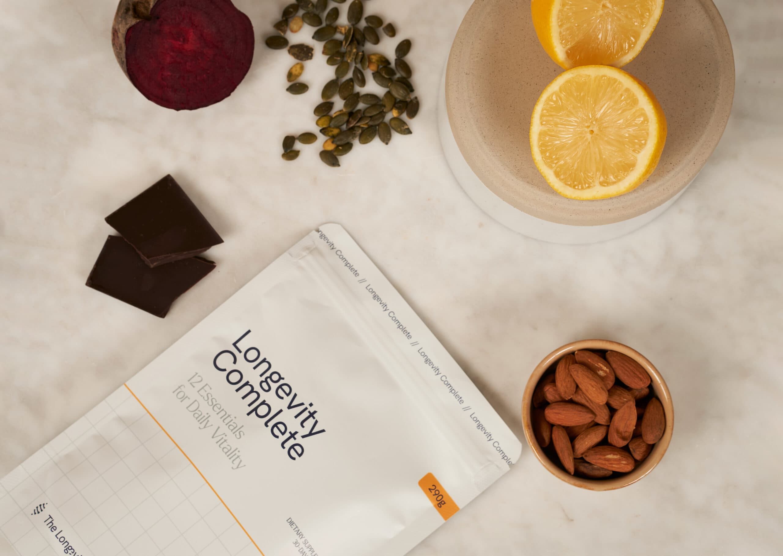 Verpackung 'Longevity Complete' mit 12 Essentials für tägliche Vitalität. Daneben Rote Bete, Kürbiskerne, Schokolade, Zitronenscheiben und Mandeln.
