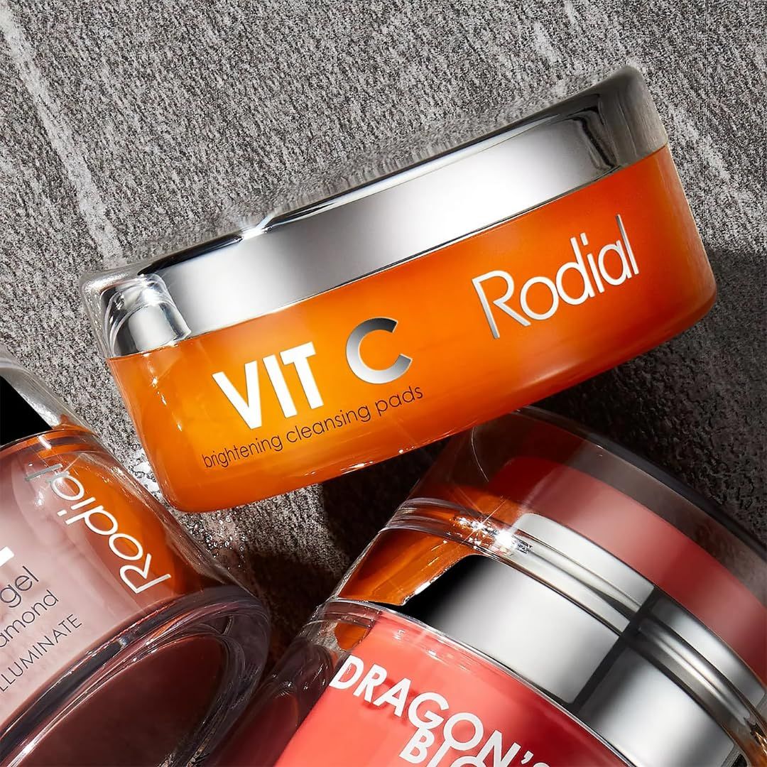 Drei Dosen auf einer Oberfläche. Aufschrift: VIT C Rodial. Daneben weitere Produkte. Orangefarbene Dose mit silbernem Deckel.